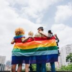 Four people stand arm in arm with their backs to the camera, holding a large rainbow Pride flag. They face a city skyline under a bright, partly cloudy sky.