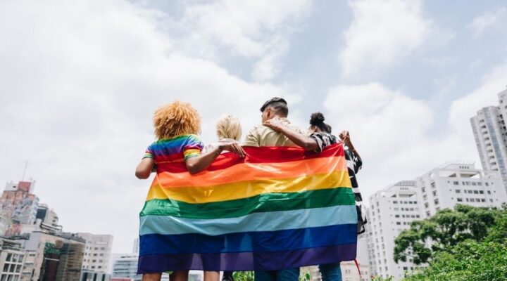 Four people stand arm in arm with their backs to the camera, holding a large rainbow Pride flag. They face a city skyline under a bright, partly cloudy sky.
