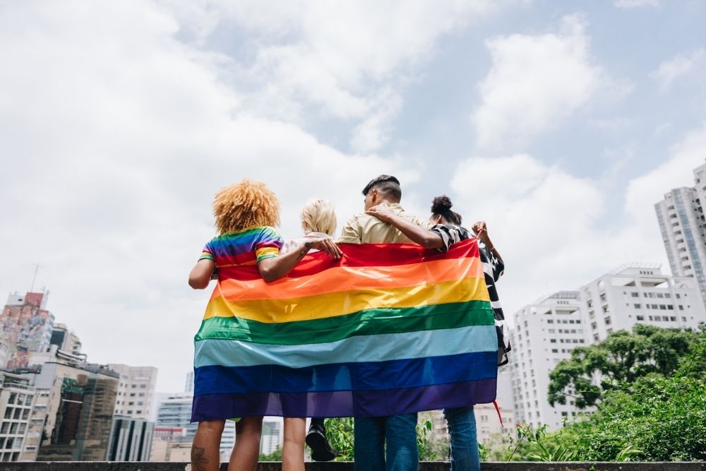 Four people stand arm in arm with their backs to the camera, holding a large rainbow Pride flag. They face a city skyline under a bright, partly cloudy sky.