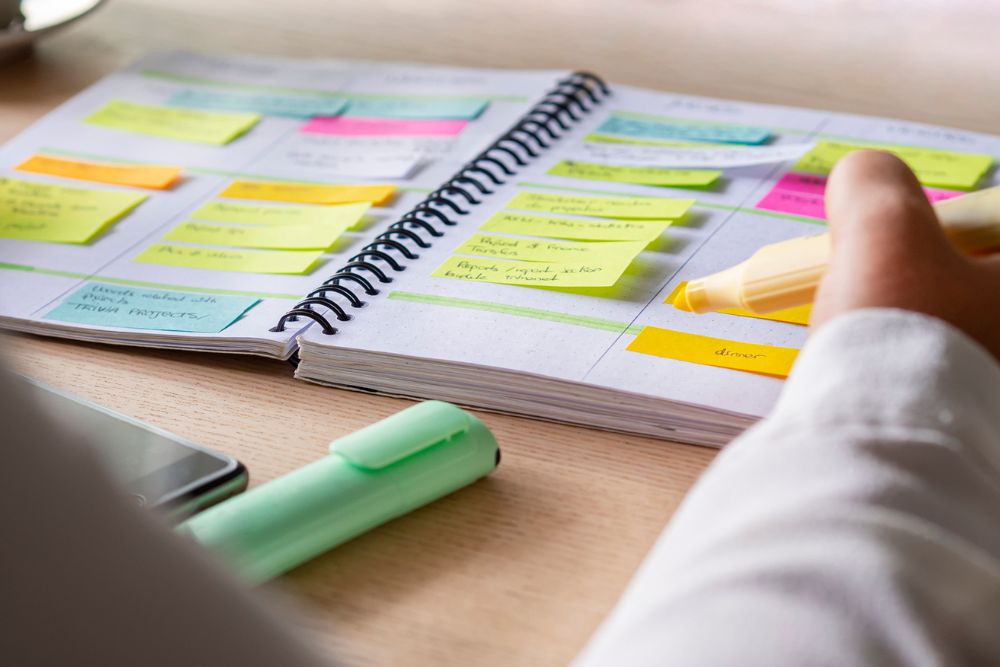 Close-up of a person highlighting tasks in a planner filled with colorful sticky notes, with a phone and a highlighter on the desk.