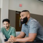 A group of men sitting in a circle, engaged in a supportive conversation, suggesting a recovery meeting or alumni support group session.