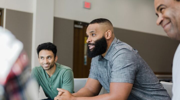 A group of men sitting in a circle, engaged in a supportive conversation, suggesting a recovery meeting or alumni support group session.