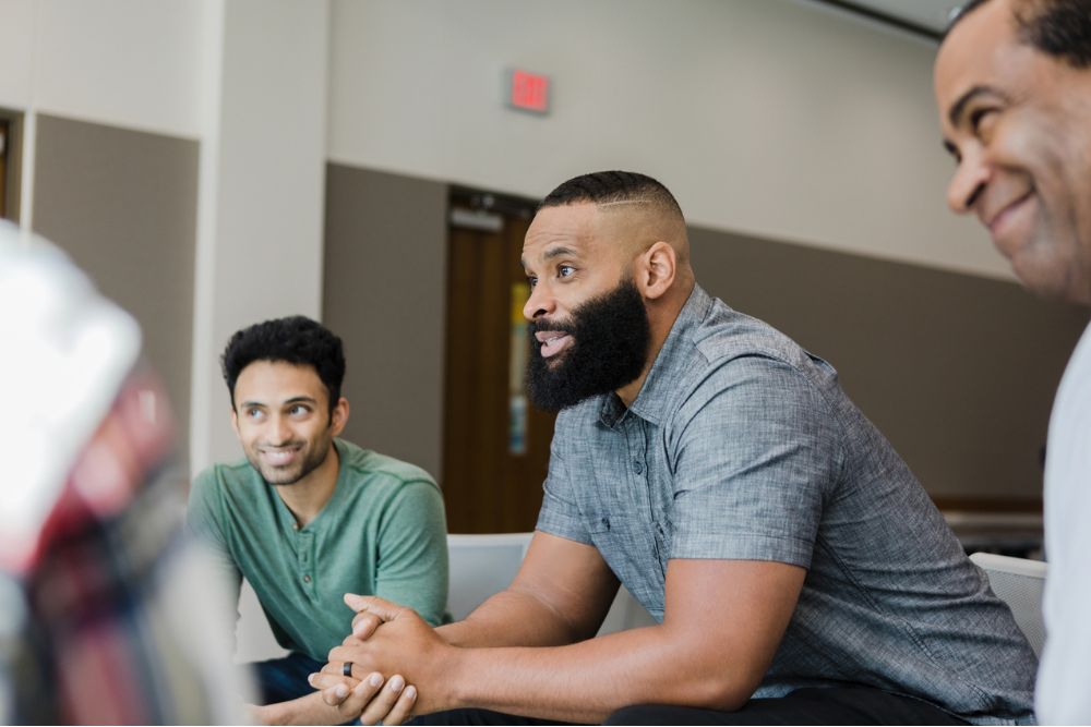 A group of men sitting in a circle, engaged in a supportive conversation, suggesting a recovery meeting or alumni support group session.