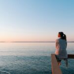 A woman sits peacefully on a wooden railing overlooking a calm lake at sunset. She gazes out over the water, wrapped in a cozy shawl.