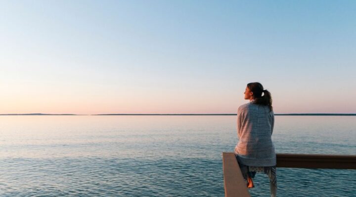 A woman sits peacefully on a wooden railing overlooking a calm lake at sunset. She gazes out over the water, wrapped in a cozy shawl.