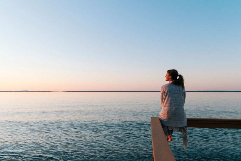 A woman sits peacefully on a wooden railing overlooking a calm lake at sunset. She gazes out over the water, wrapped in a cozy shawl.