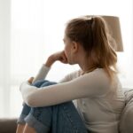 Young woman sitting on a couch with her knees pulled to her chest, looking out a bright window with a pensive expression, symbolizing reflection or emotional struggle.