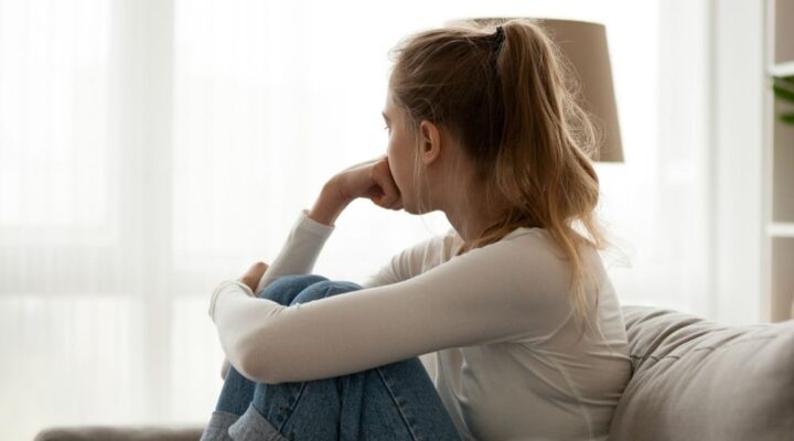 Young woman sitting on a couch with her knees pulled to her chest, looking out a bright window with a pensive expression, symbolizing reflection or emotional struggle.