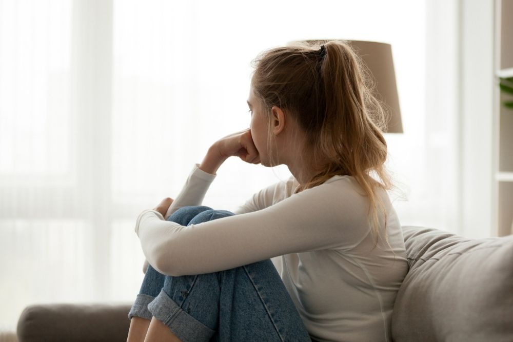 Young woman sitting on a couch with her knees pulled to her chest, looking out a bright window with a pensive expression, symbolizing reflection or emotional struggle.