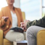 Close-up of a therapy session with two people seated in armchairs, one person speaking and the other listening with hands clasped, suggesting counseling or cognitive behavioral therapy.