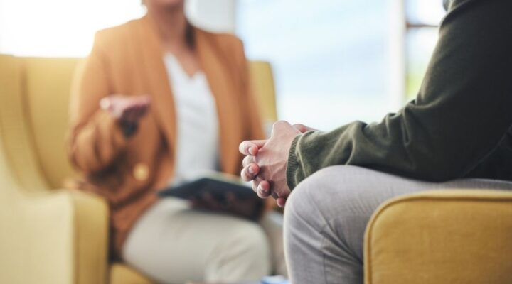 Close-up of a therapy session with two people seated in armchairs, one person speaking and the other listening with hands clasped, suggesting counseling or cognitive behavioral therapy.