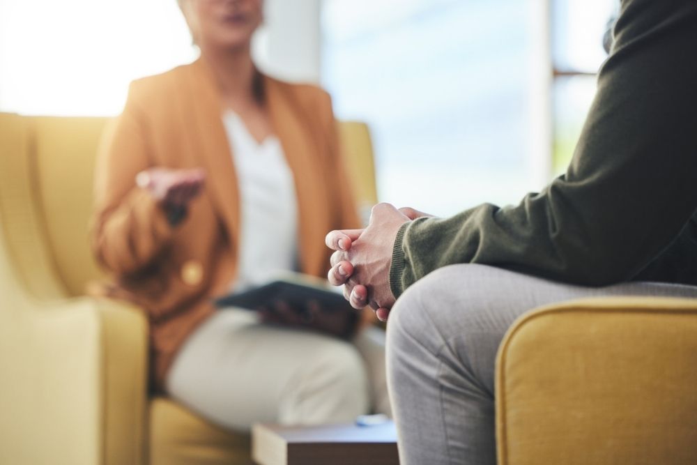 Close-up of a therapy session with two people seated in armchairs, one person speaking and the other listening with hands clasped, suggesting counseling or cognitive behavioral therapy.