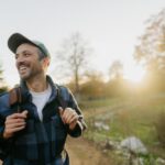 A smiling man with a backpack walks outdoors on a sunny day, enjoying nature with trees and a glowing sunset in the background.