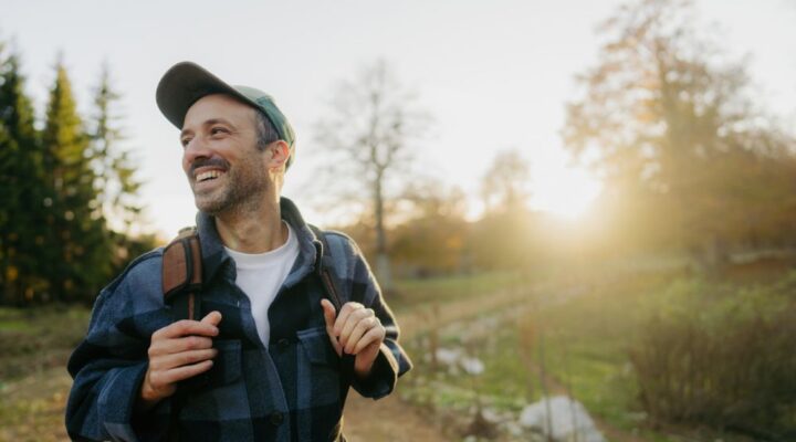 A smiling man with a backpack walks outdoors on a sunny day, enjoying nature with trees and a glowing sunset in the background.