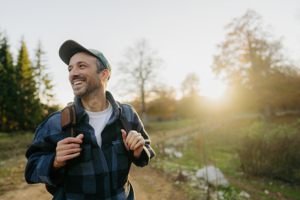 A smiling man with a backpack walks outdoors on a sunny day, enjoying nature with trees and a glowing sunset in the background.