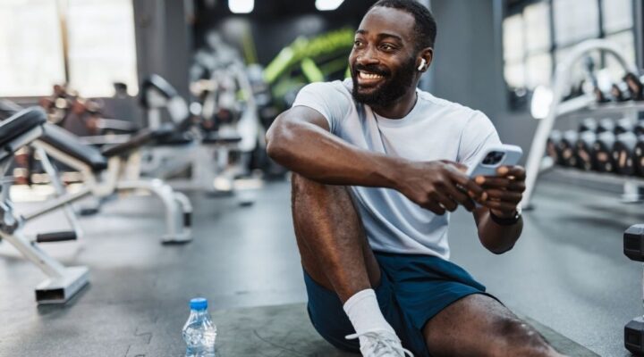 Man sitting on the floor of a gym smiling, holding a phone and water bottle, taking a break from working out.