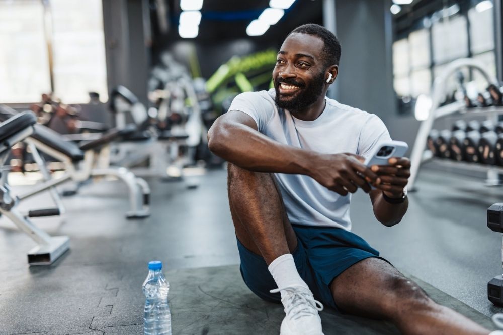 Man sitting on the floor of a gym smiling, holding a phone and water bottle, taking a break from working out.