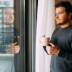 A man stands by a window holding a coffee mug, looking outside thoughtfully
