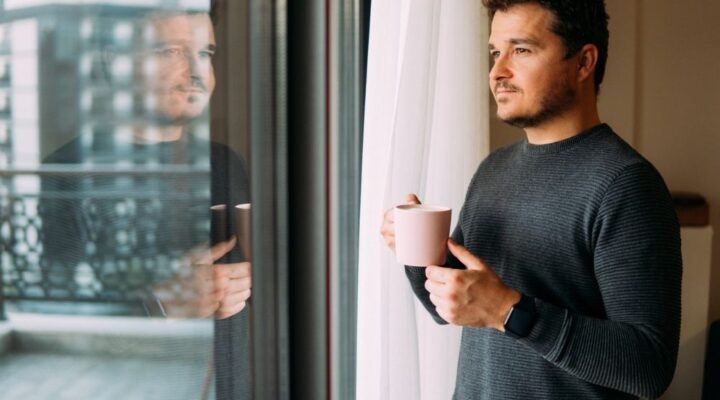 A man stands by a window holding a coffee mug, looking outside thoughtfully