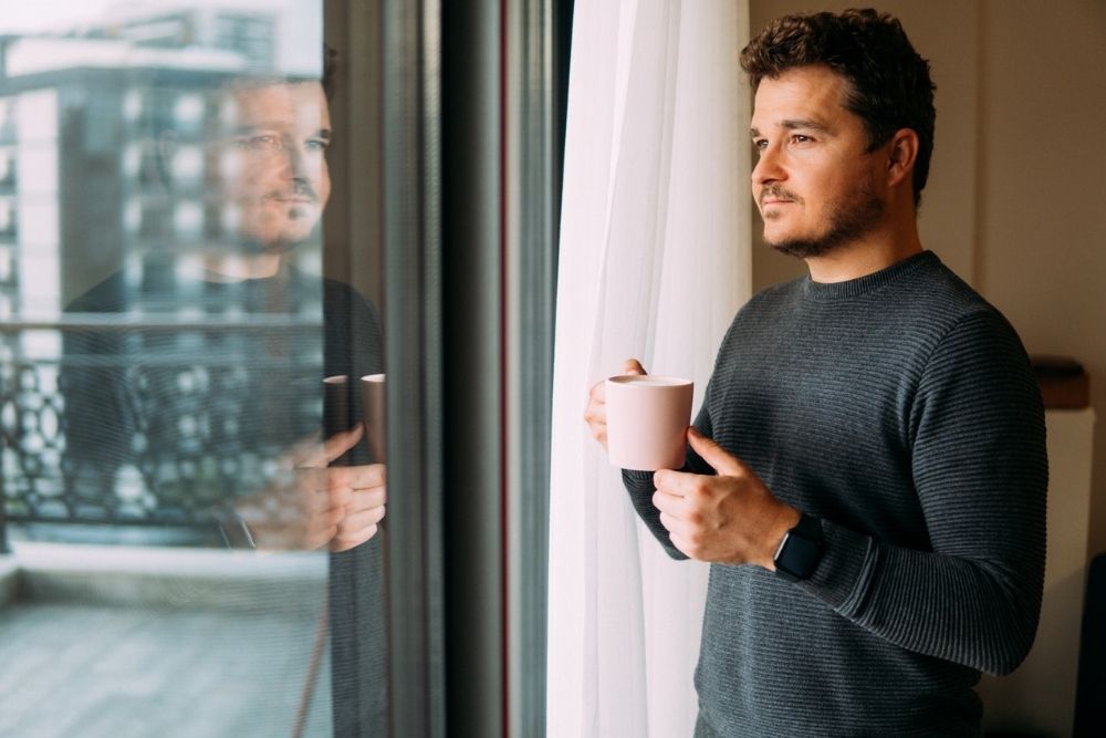 A man stands by a window holding a coffee mug, looking outside thoughtfully