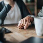 Close-up of two people sitting at a table holding hands in a supportive gesture, with coffee cups nearby.