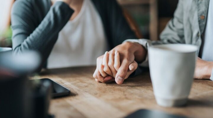Close-up of two people sitting at a table holding hands in a supportive gesture, with coffee cups nearby.