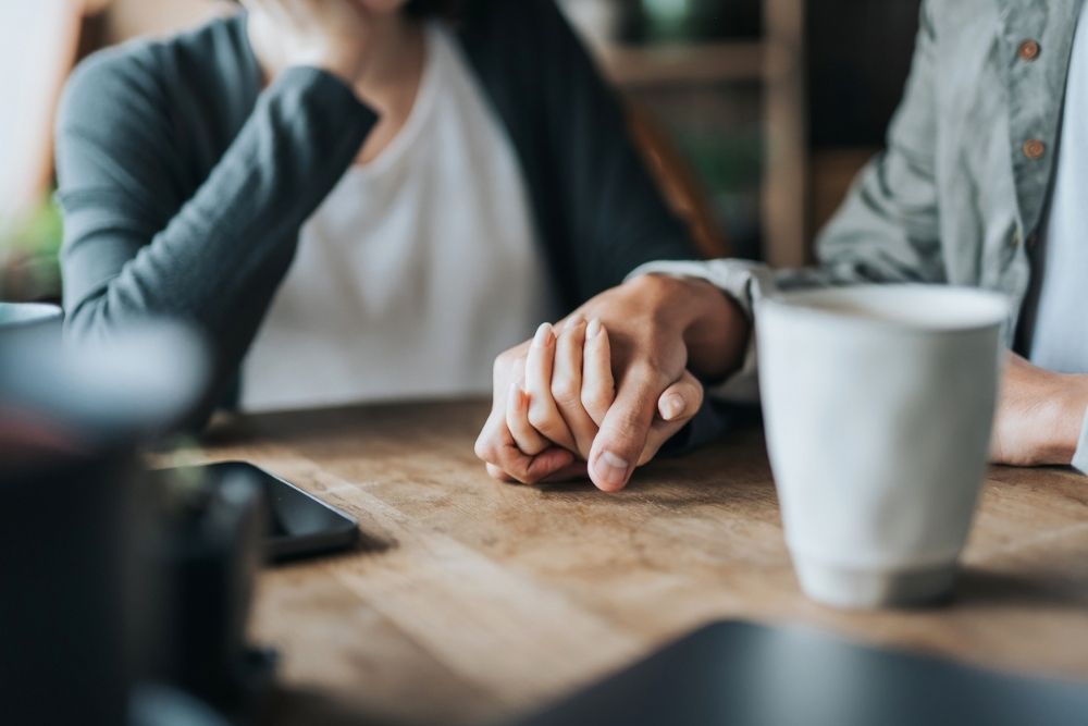Close-up of two people sitting at a table holding hands in a supportive gesture, with coffee cups nearby.