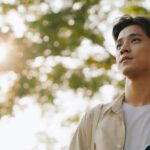 Young man standing outdoors with a calm, thoughtful expression, sunlight filtering through trees in the background.