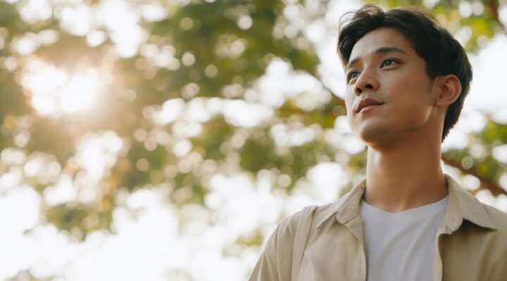Young man standing outdoors with a calm, thoughtful expression, sunlight filtering through trees in the background.