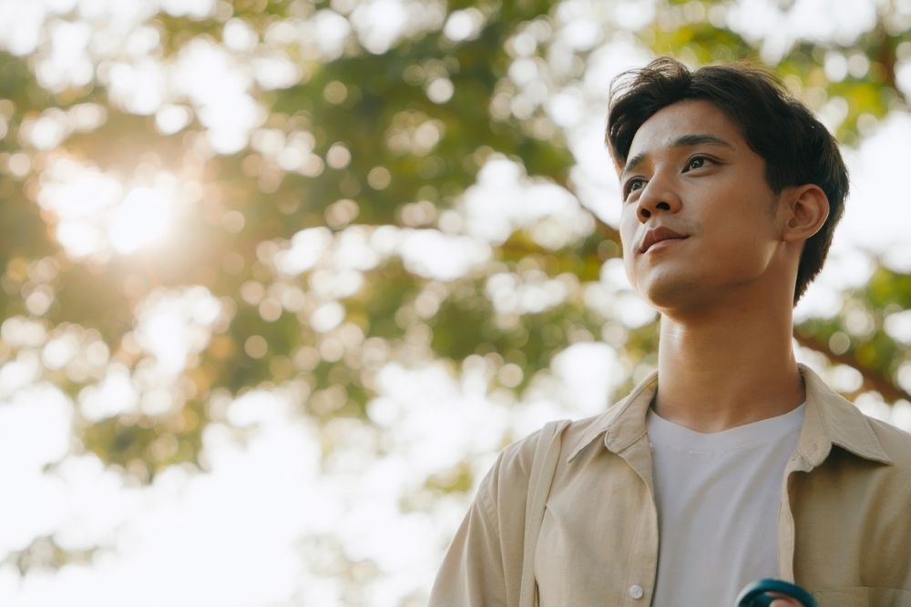 Young man standing outdoors with a calm, thoughtful expression, sunlight filtering through trees in the background.