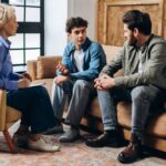 A teenage boy sits between two adults during a family therapy session, all engaged in conversation on a living room-style couch.