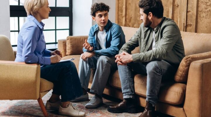 A teenage boy sits between two adults during a family therapy session, all engaged in conversation on a living room-style couch.