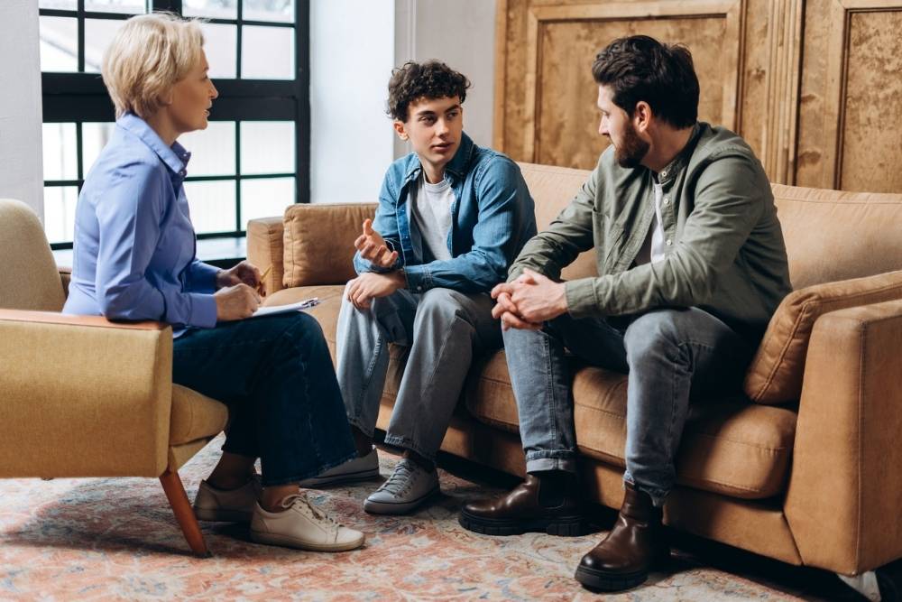 A teenage boy sits between two adults during a family therapy session, all engaged in conversation on a living room-style couch.