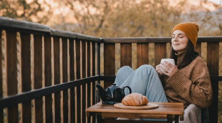 A woman sits on a wooden balcony in cool weather, wearing a beanie and cozy sweater. With her eyes closed, she holds a warm drink and relaxes next to a small table with tea and a pastry, enjoying a peaceful autumn morning.