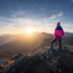 A person stands on a mountain overlook at sunrise, facing the horizon with a sense of reflection, strength, and calm.