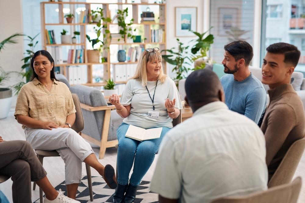 A group of people sitting in a circle during a therapy session, with a facilitator speaking and participants engaged and listening.