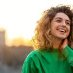 A smiling person outdoors, looking upward with a relaxed expression as sunlight shines behind them.