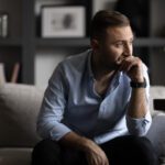 A man sits on a couch with his hand to his face, looking thoughtful and concerned in a quiet living room.