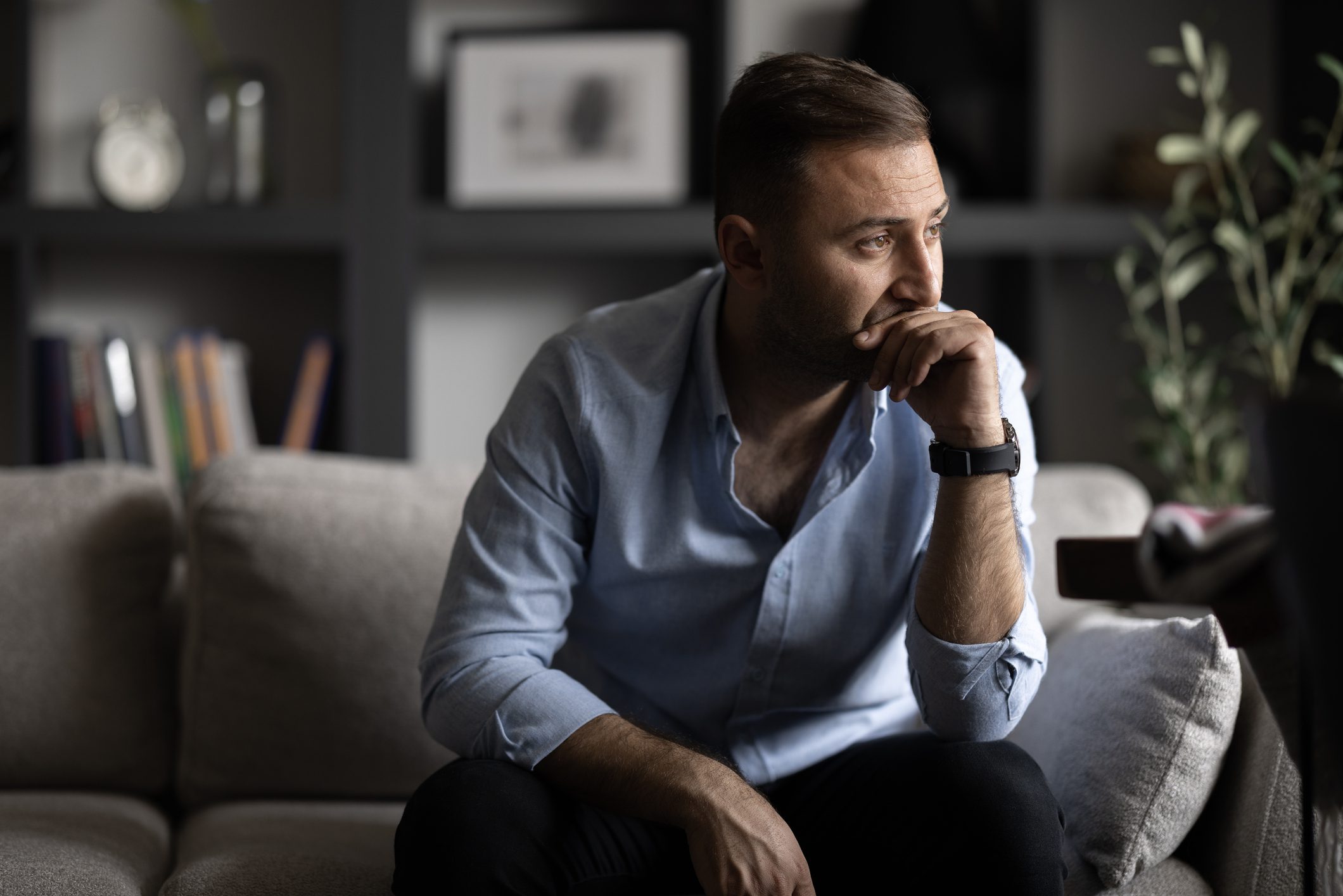 A man sits on a couch with his hand to his face, looking thoughtful and concerned in a quiet living room.