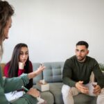 A couple sits on a sofa during a counseling session, appearing engaged and thoughtful as they explore ways to approach conflict. A counselor with a notebook listens attentively, creating a supportive atmosphere.