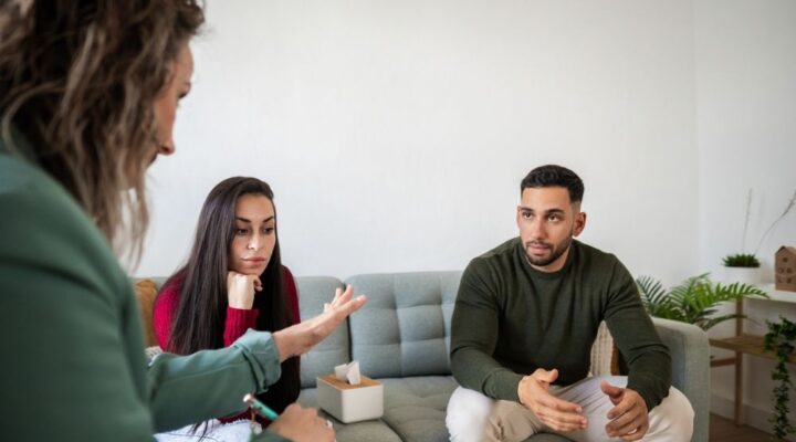 A couple sits on a sofa during a counseling session, appearing engaged and thoughtful as they explore ways to approach conflict. A counselor with a notebook listens attentively, creating a supportive atmosphere.