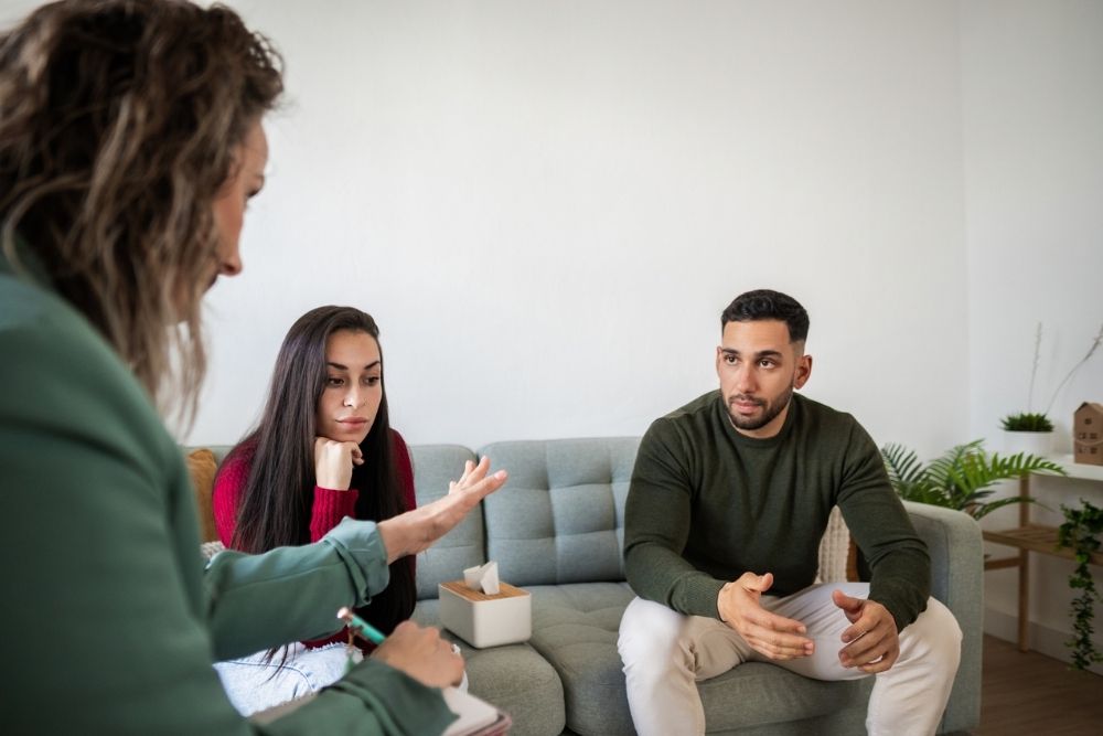 A couple sits on a sofa during a counseling session, appearing engaged and thoughtful as they explore ways to approach conflict. A counselor with a notebook listens attentively, creating a supportive atmosphere.