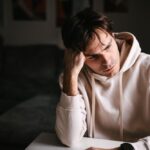 Man in a white hoodie sits indoors with his head resting on his hand, looking stressed and exhausted.