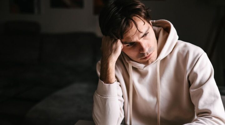 Man in a white hoodie sits indoors with his head resting on his hand, looking stressed and exhausted.