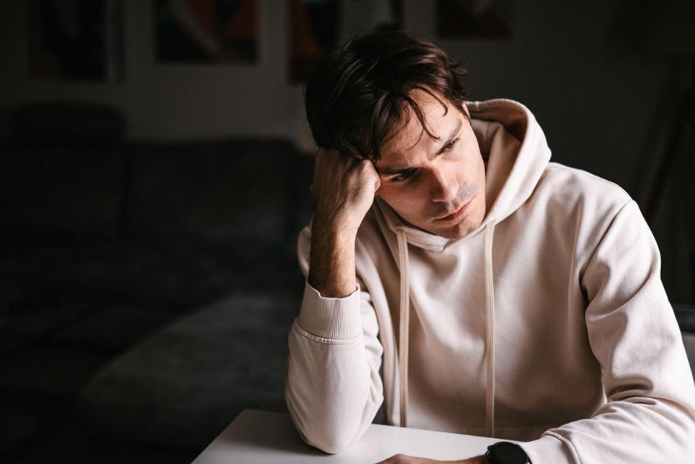 Man in a white hoodie sits indoors with his head resting on his hand, looking stressed and exhausted.