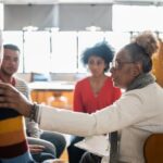 Ivory Plains - Stand Firm When Seeking LGBTQ+ Affirming Care. A group of individuals sits in a circle. An older woman places her hand on a mans shoulder as he shares with the group.