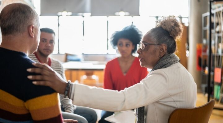 Ivory Plains - Stand Firm When Seeking LGBTQ+ Affirming Care. A group of individuals sits in a circle. An older woman places her hand on a mans shoulder as he shares with the group.