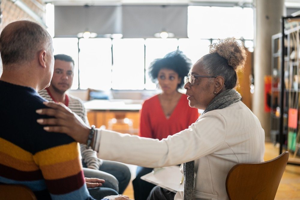 Ivory Plains - Stand Firm When Seeking LGBTQ+ Affirming Care. A group of individuals sits in a circle. An older woman places her hand on a mans shoulder as he shares with the group.